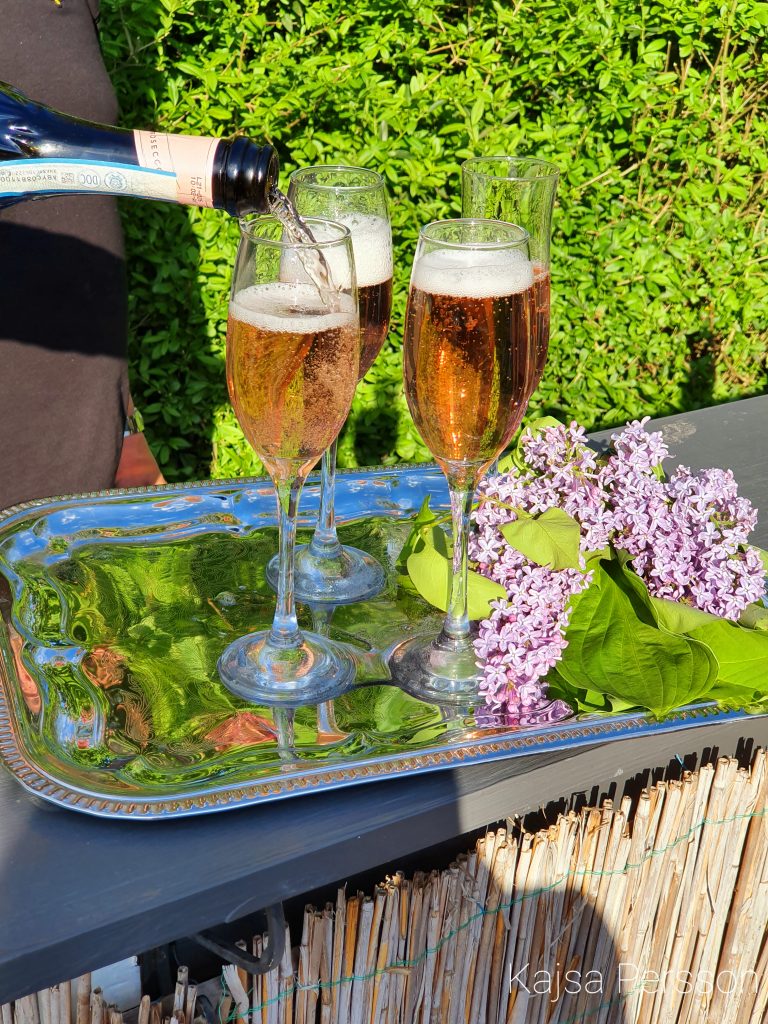 Fyra Champagneglas där man häller upp rosé champagne. silverbricka med syrenblommor